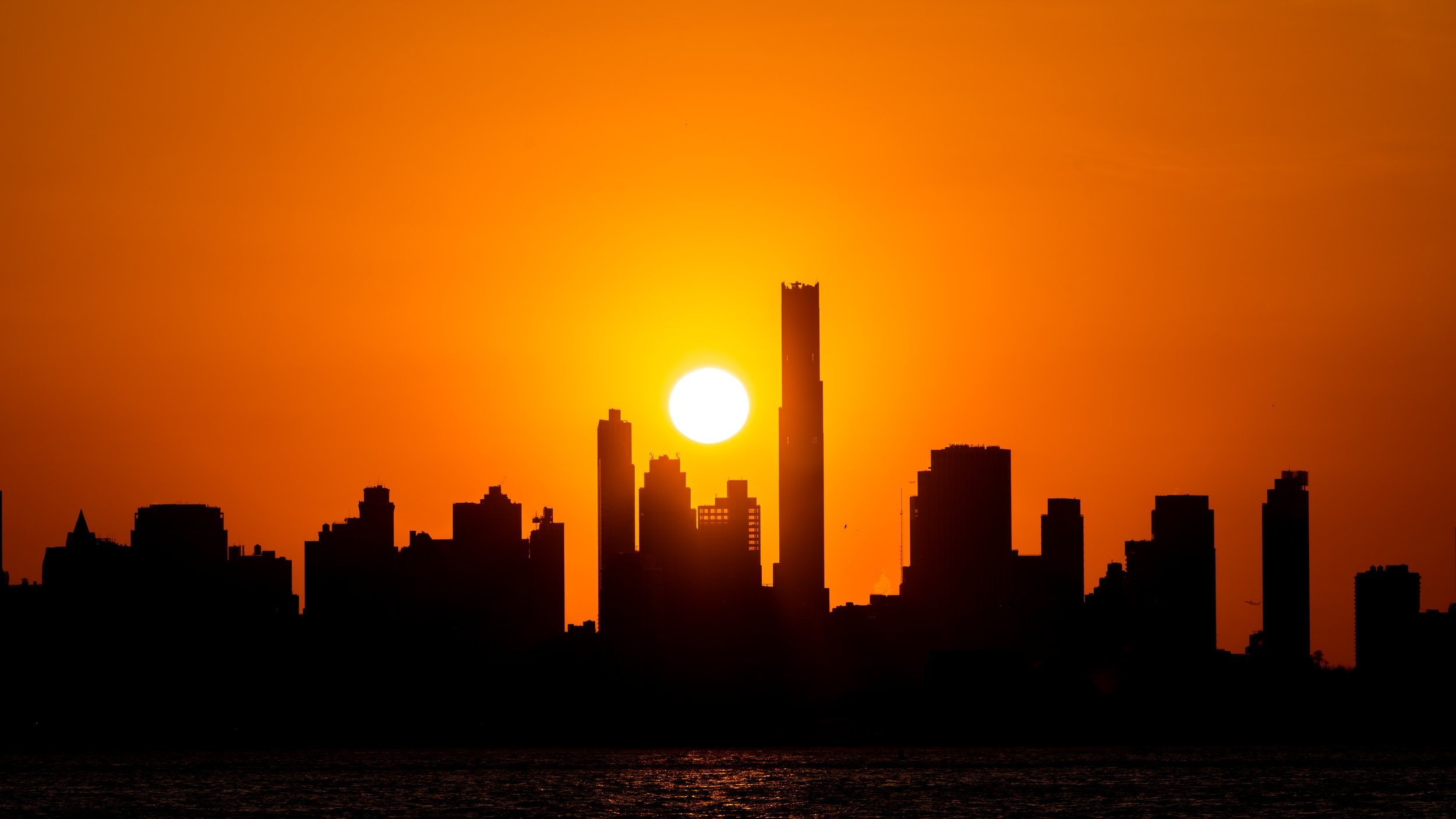 Sunset over NYC skyline with tall buildings silhouetted against an orange sky and reflection on water.