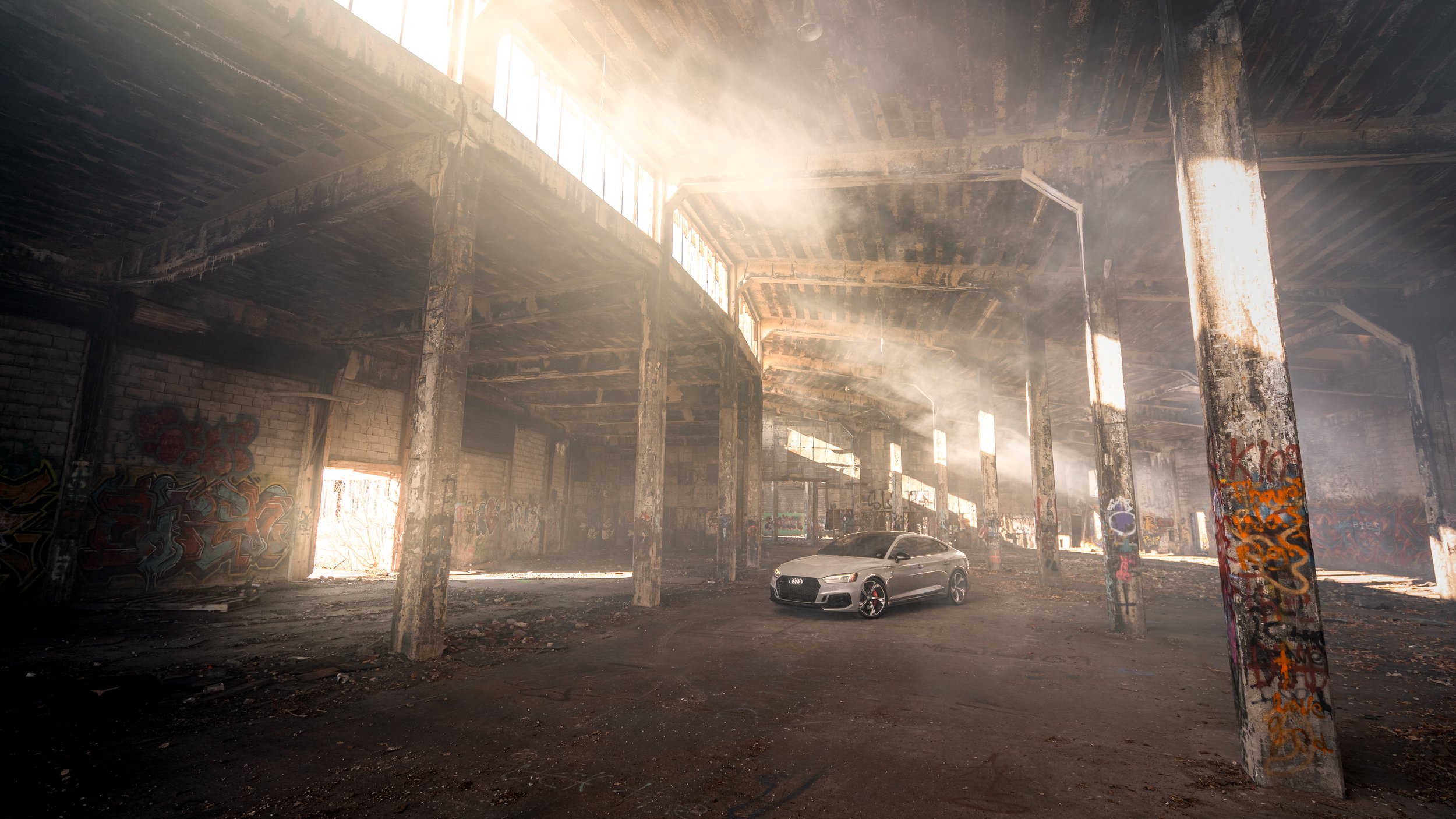 An Audi RS7 parked inside an abandoned, graffiti-covered warehouse with beams and windows, sunlight streaming in.