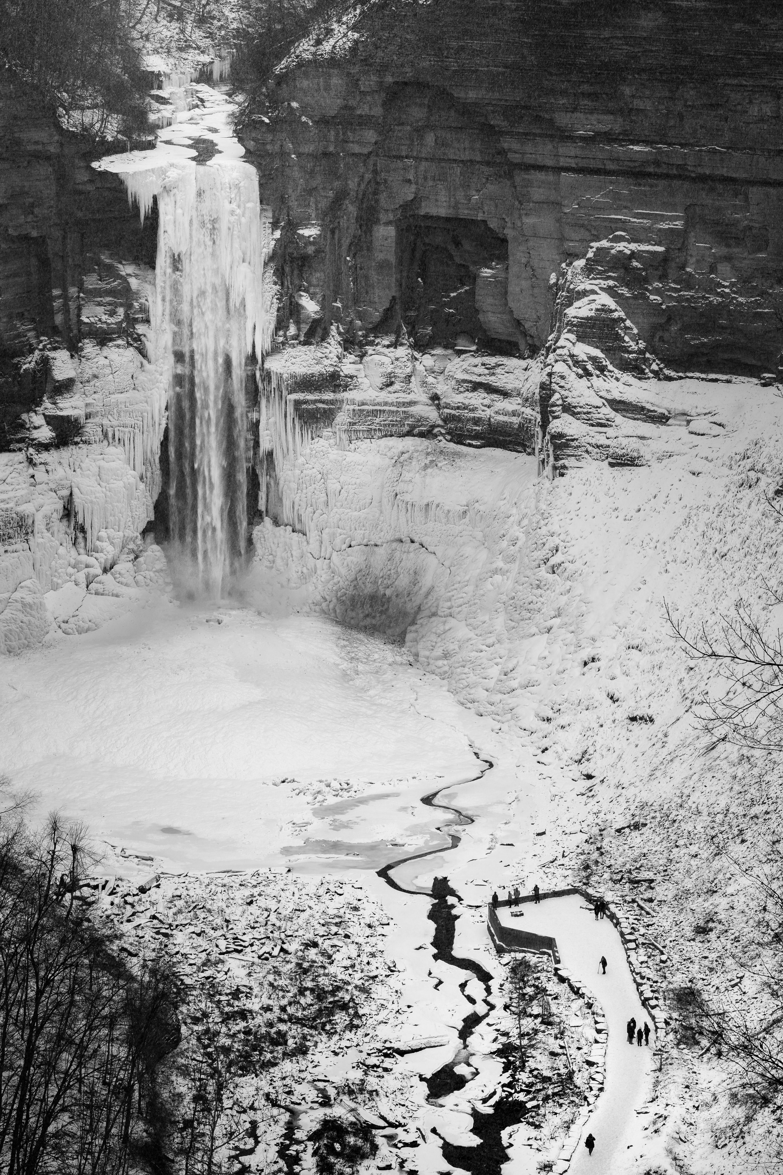 Snow-covered landscape of Ithaca with a frozen waterfall, icy river, and a viewing platform with visitors.
