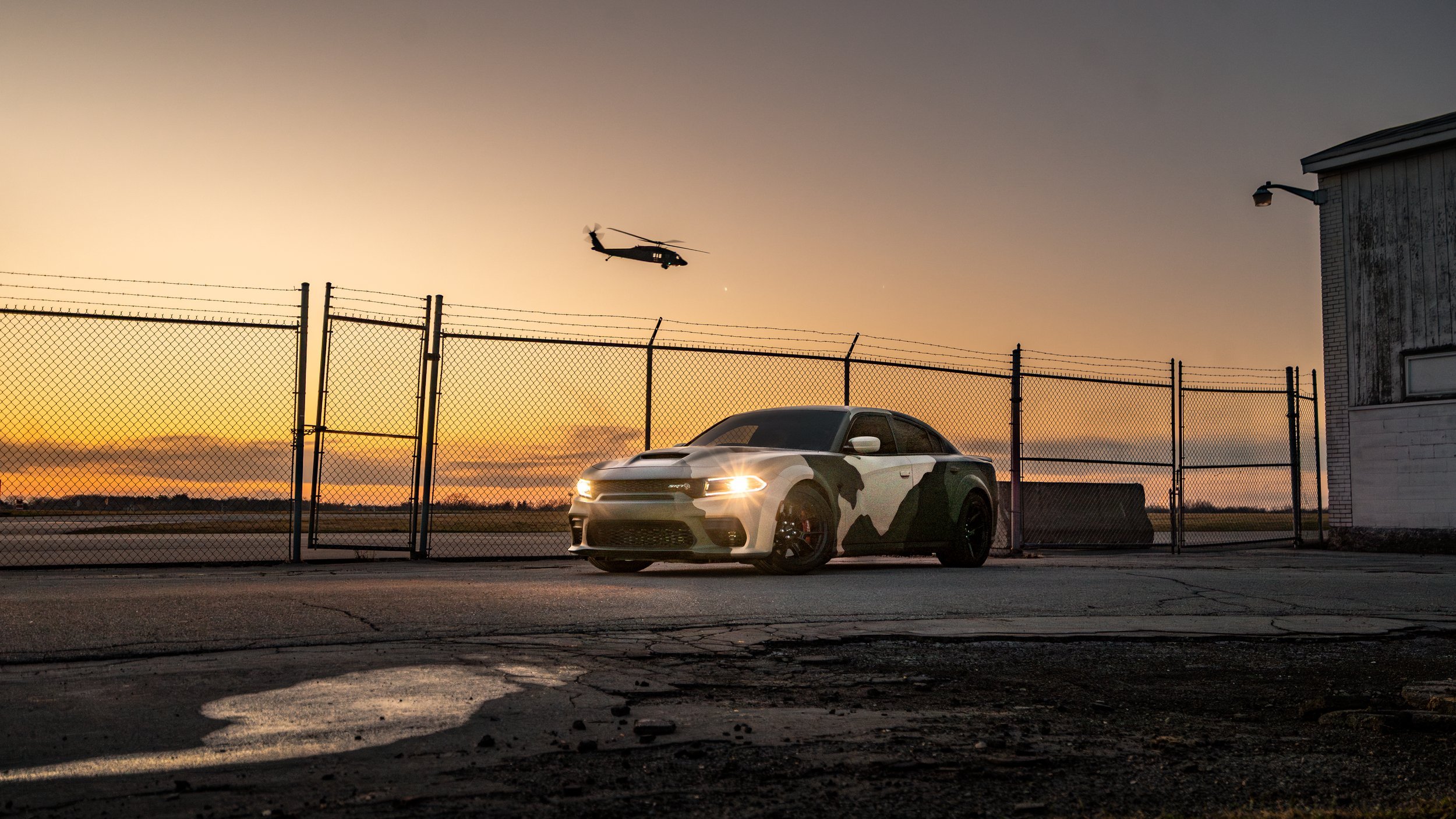 Camo Dodge Charger SRT parked behind a chain-link fence at sunset, with a black hawk military helicopter flying in the sky nearby.