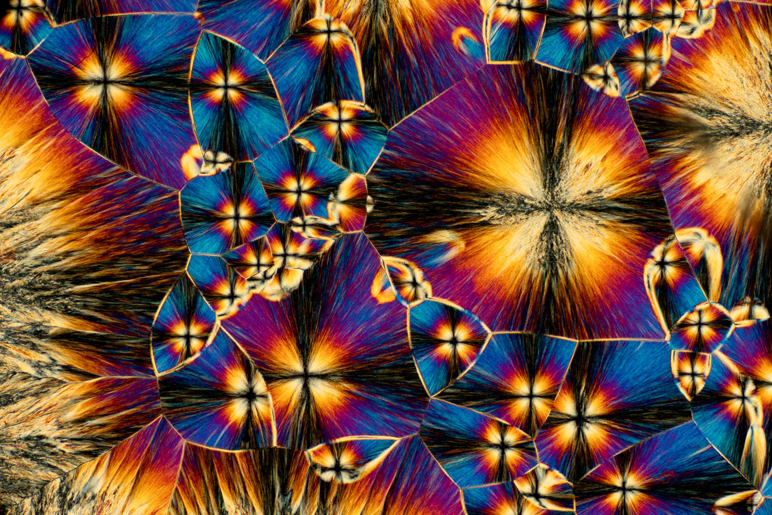 Microscopic cross-polarization of a crystal with vibrant, multicolored patterns in shades of blue, orange, purple, and black, displaying a fractured, layered appearance.