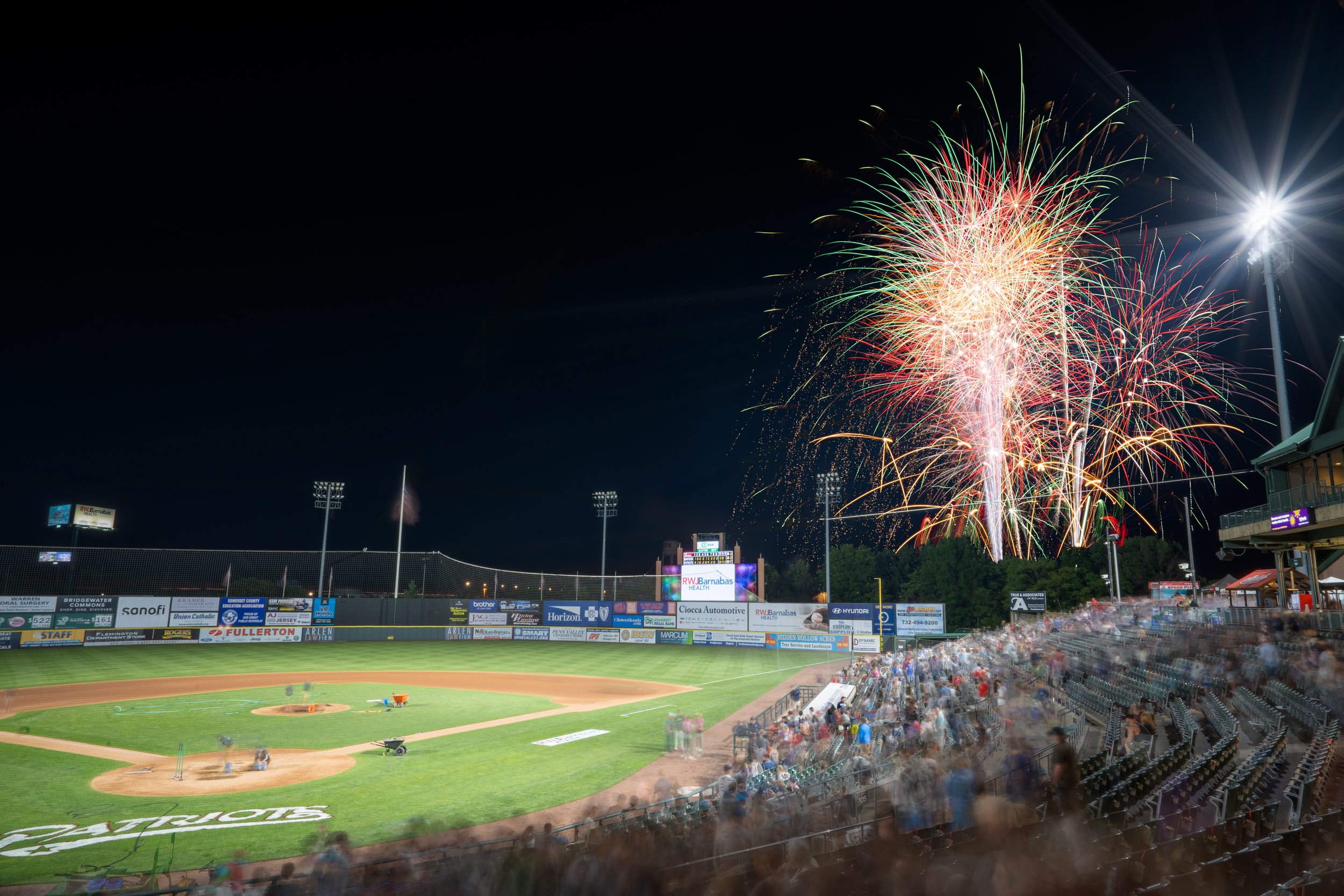 Fireworks display over a baseball stadium at night, with spectators in the stands and advertisements on the outfield wall.