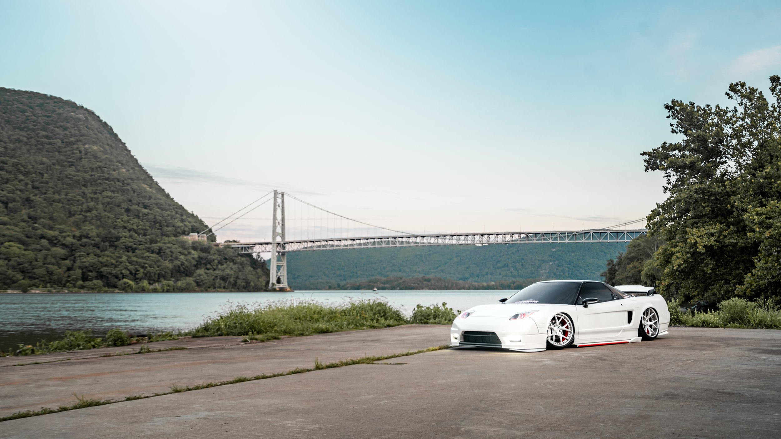 A white sports car with black tinted windows parked on a concrete surface near a large body of water with a bridge in the background, surrounded by green hills and trees.