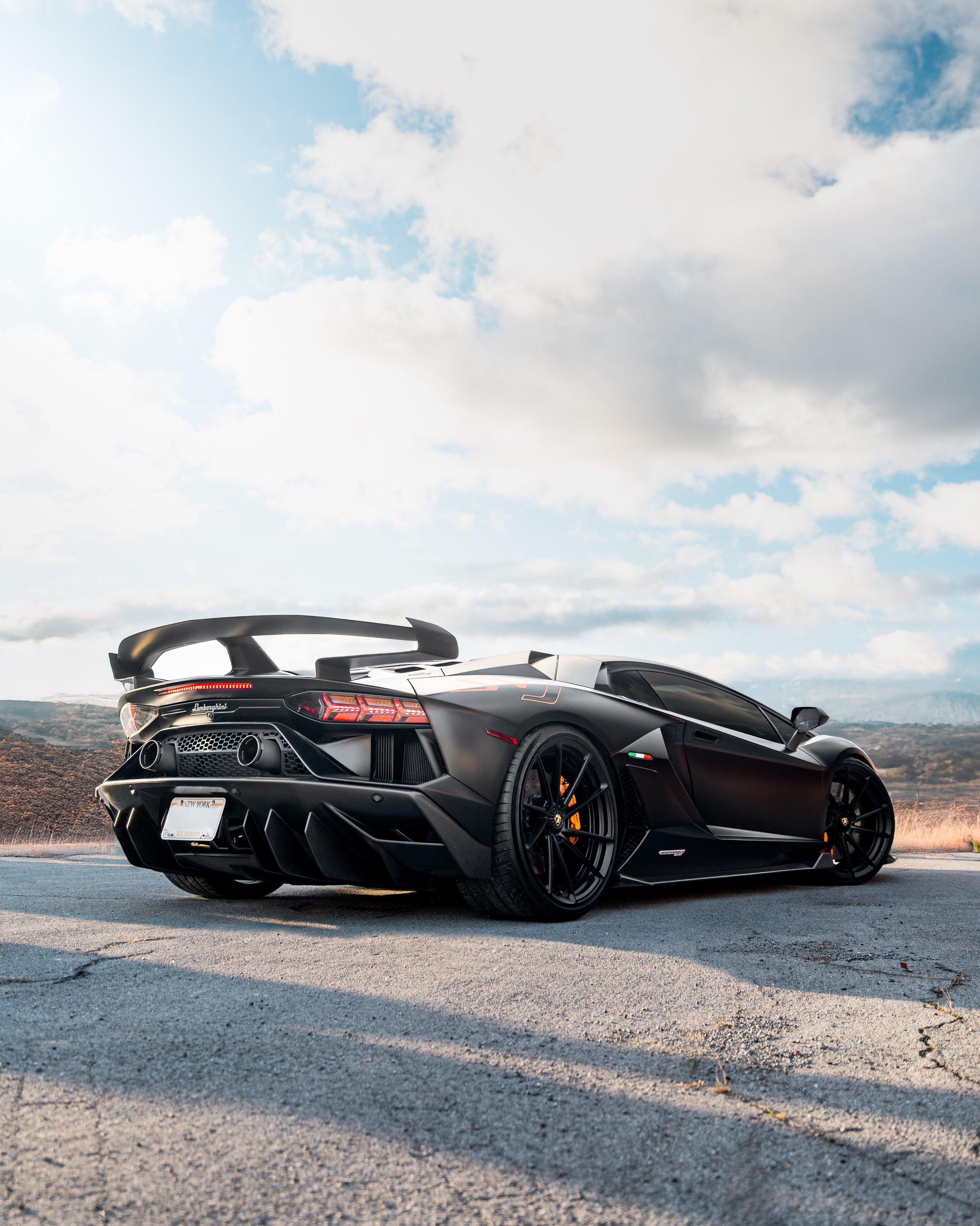 Black Lamborghini SVJ parked on a cracked pavement with a mountainous landscape and partly cloudy sky in the background.