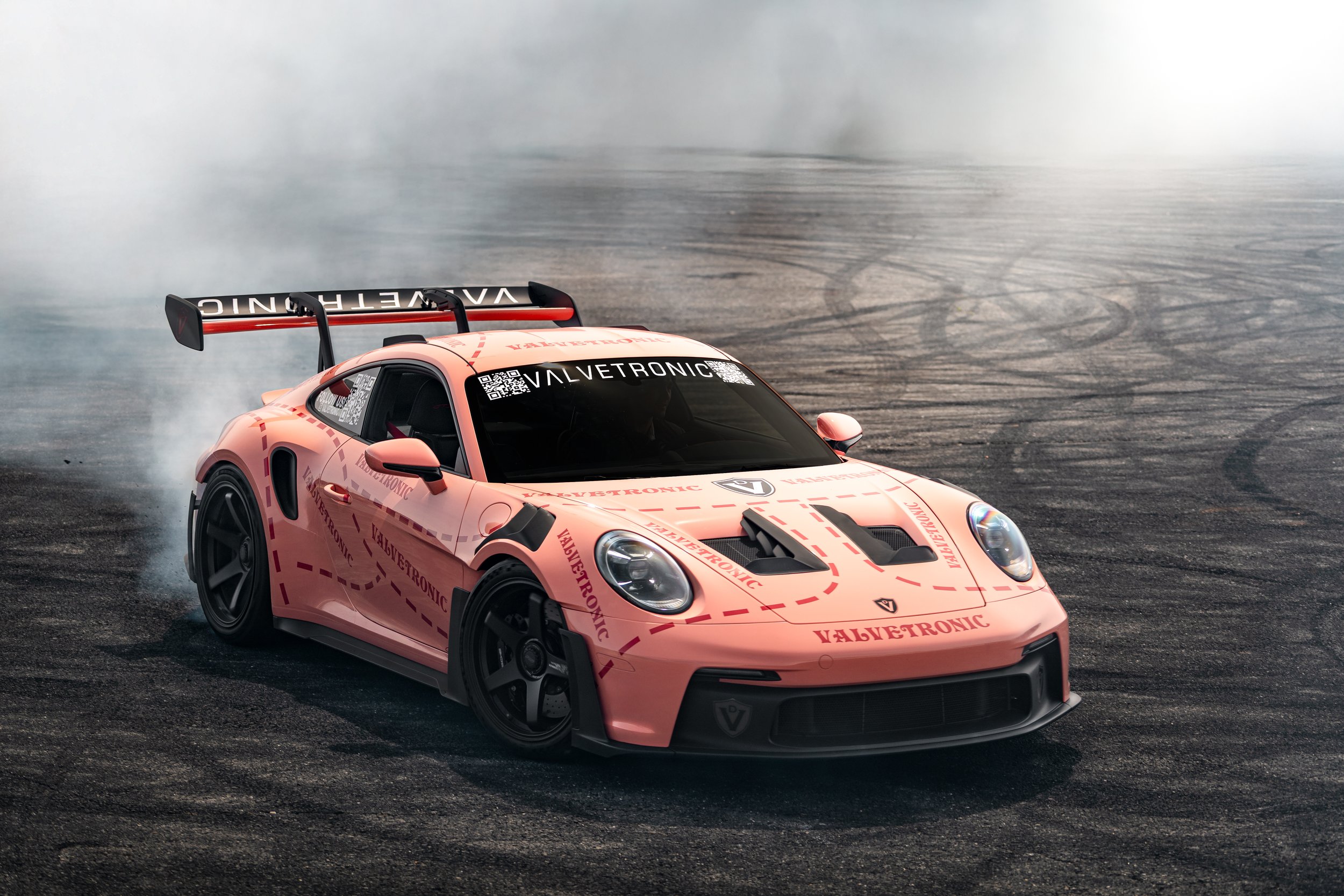 Valvetronics Porsche GT3rs drifting on a track, kicking up smoke, with VALVETRONIC branding on its hood and windshield, and a black rear wing.