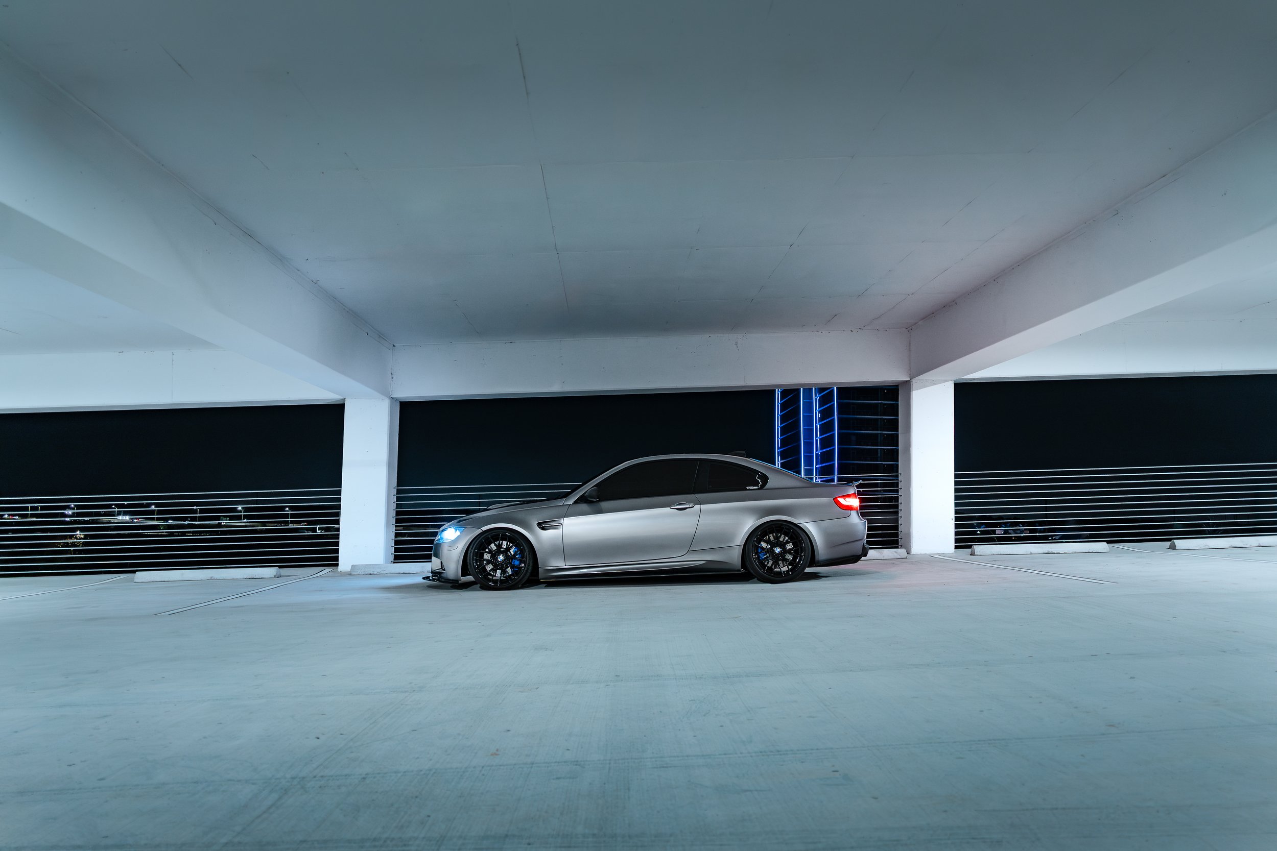 A silver sports car with tinted windows parked in an empty, multi-level parking garage at night, illuminated by nearby city lights and blue neon accents.