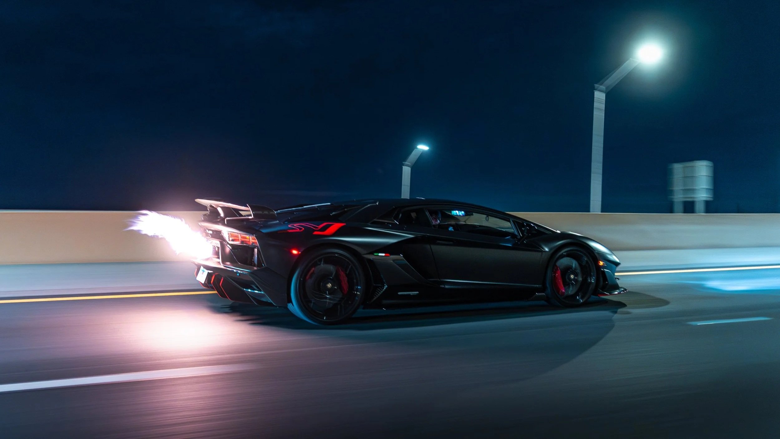 A black Lamborghini SVJ with red accents features a lit exhaust, driving at night on a highway under streetlights.