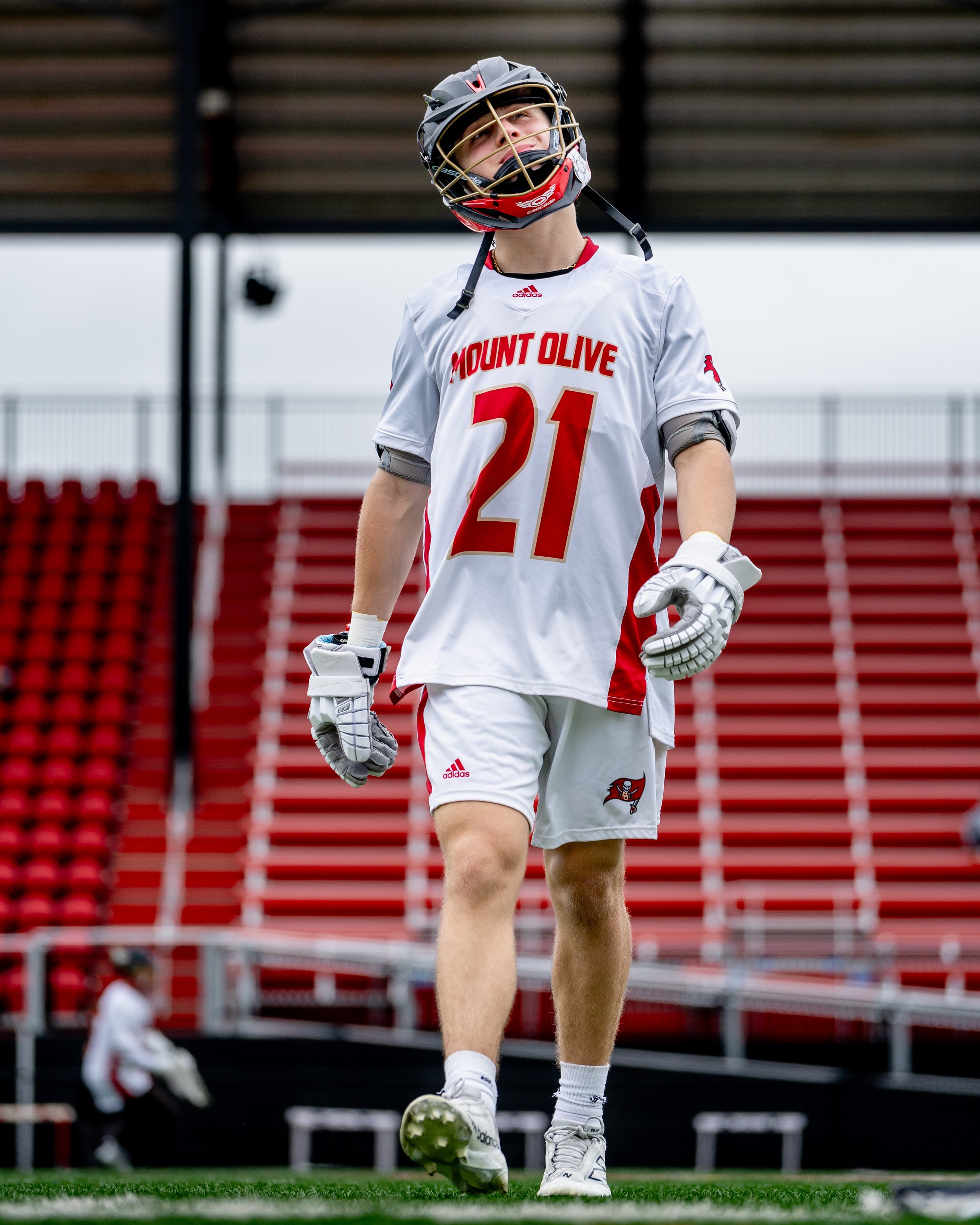 A young male lacrosse goalie in Mount Olive uniform standing on a field wearing a helmet, gloves, and athletic gear.