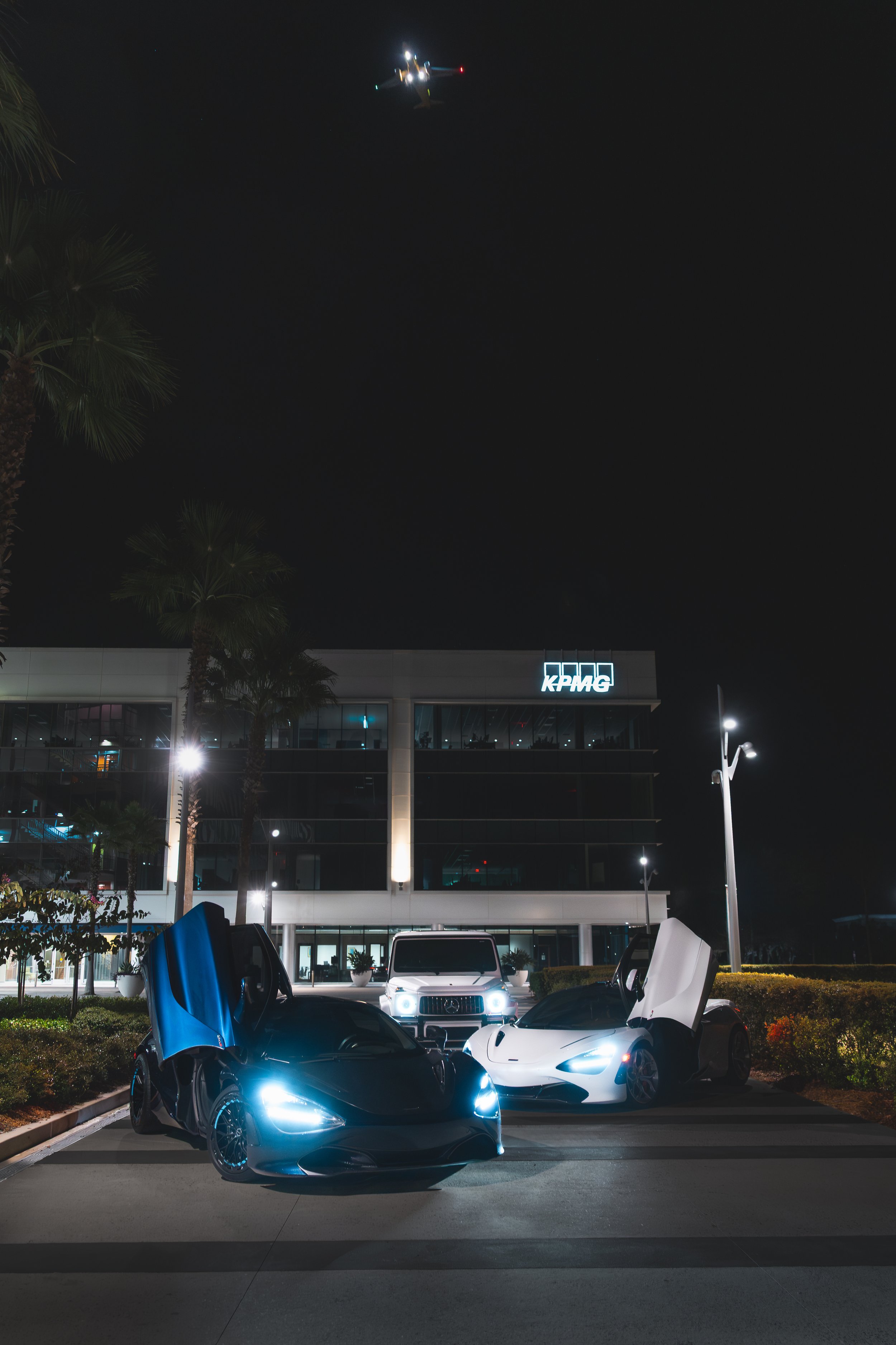 Nighttime scene featuring two luxury sports cars with vertically opening doors, a white SUV, and a modern office building with a KPMG logo, palm trees, and a helicopter flying above.