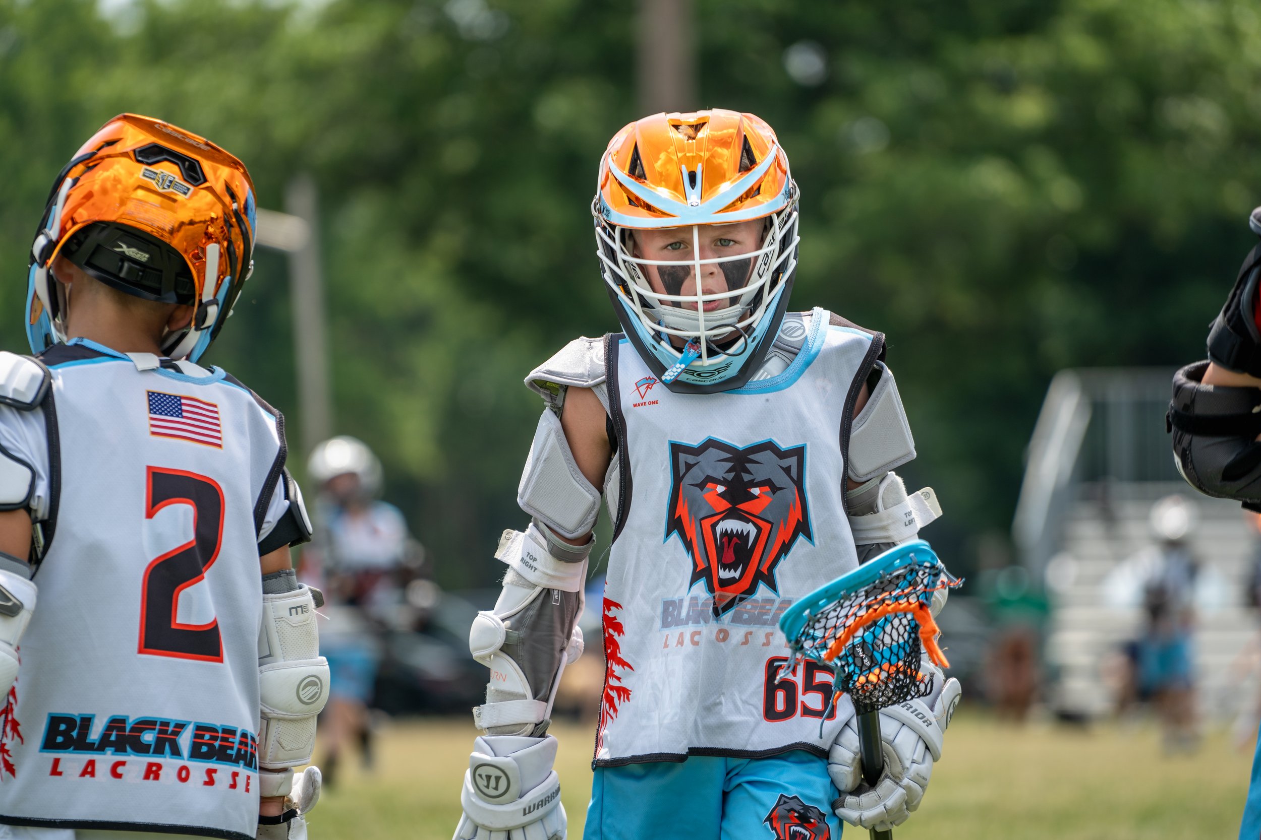 Youth lacrosse players wearing helmets and jerseys, with one player holding a lacrosse stick, on a grassy field with trees in the background.