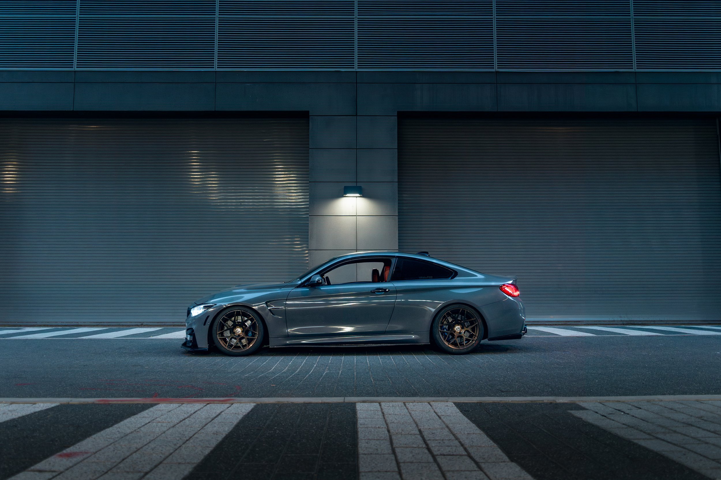 A BMW F82 parked in front of a building with a closed metal garage door at night, illuminated by wall-mounted lights in Tampa
