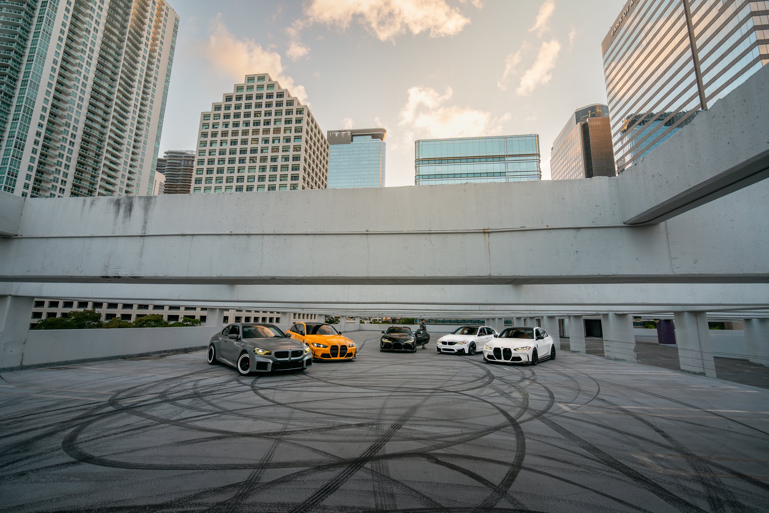 Five luxury sports cars parked on a rooftop parking garage with a city skyline of tall buildings in the background in Miami