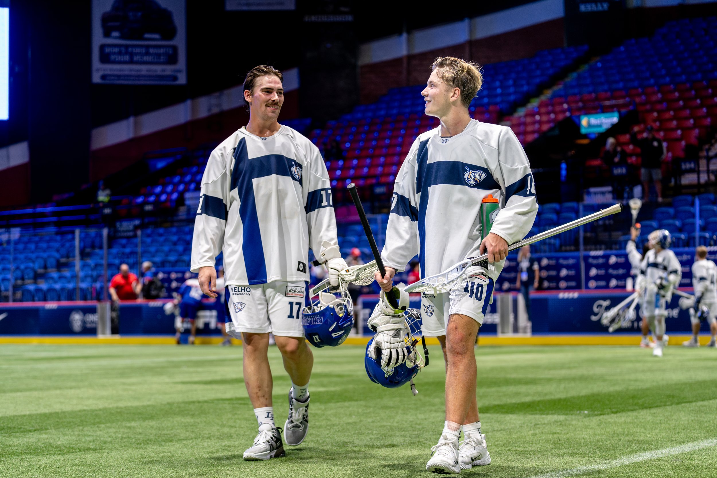Two male lacrosse players from Team Finland walking and talking on a field inside a stadium, holding lacrosse sticks and helmets at the World Box Lacrosse Tournament in Utica, NY