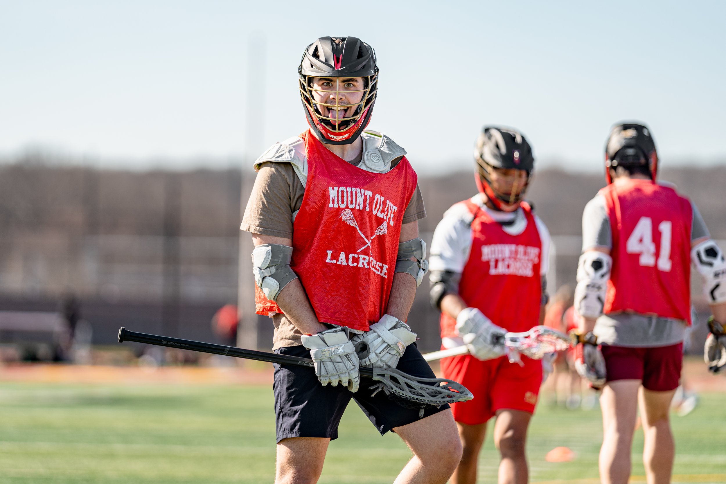 Mount Olive High School Boys Lacrosse practicing on a field, one player in the foreground is smiling and holding a lacrosse stick.