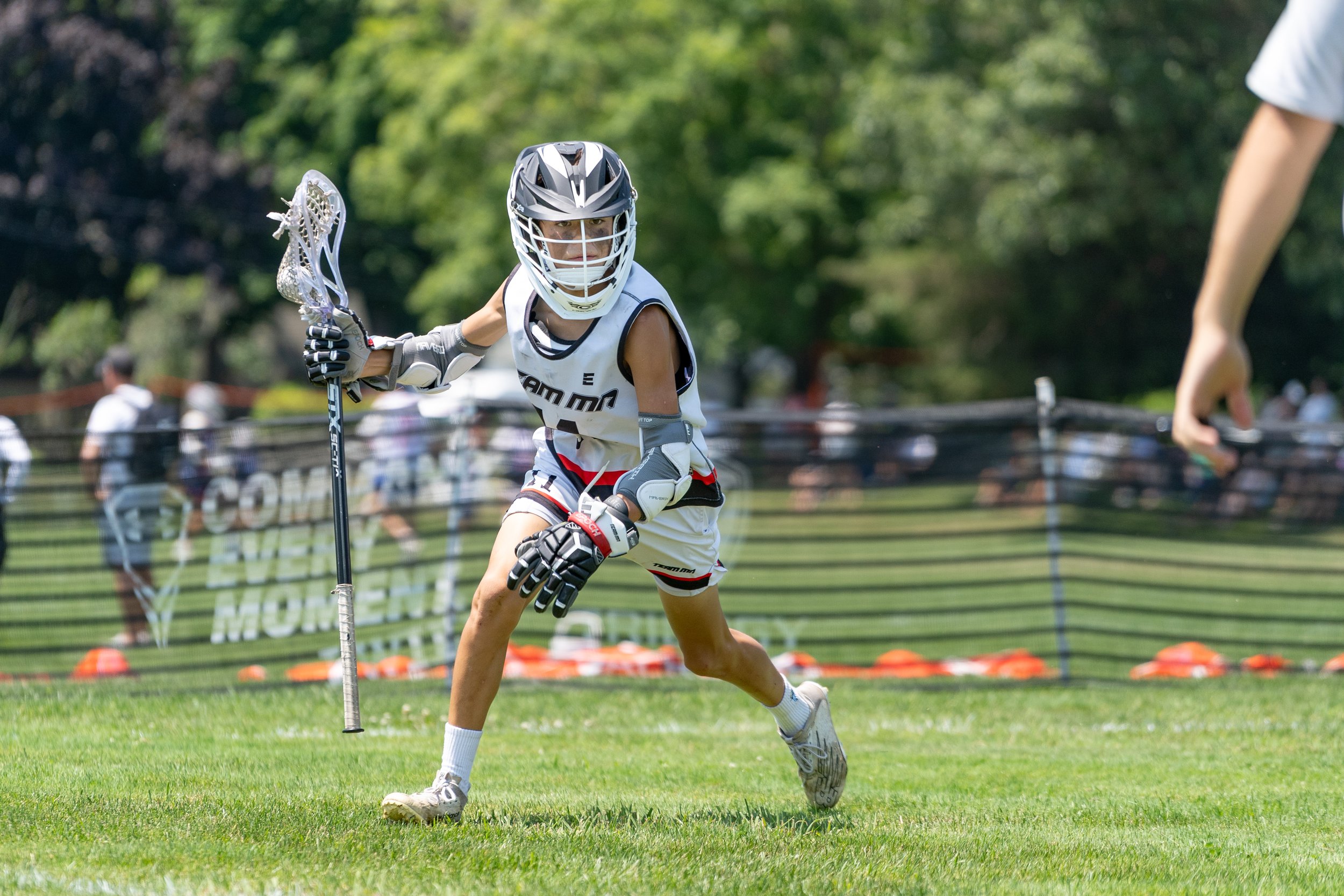 A young lacrosse player in a white uniform, helmet, and gloves, is running on a grassy field while holding a lacrosse stick. There are trees and spectators in the background.