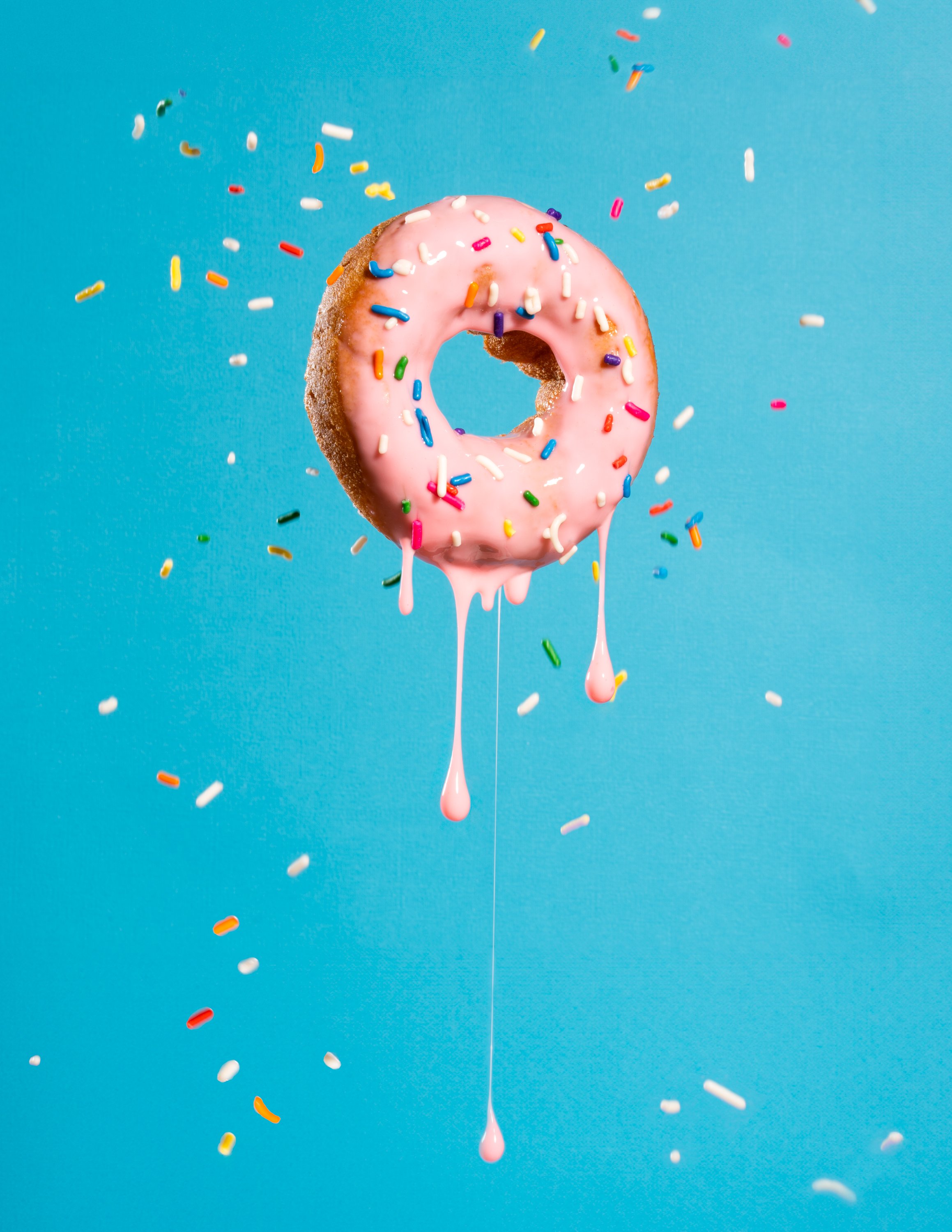 Pink-frosted donut with colorful sprinkles falling and dripping icing against a blue background.